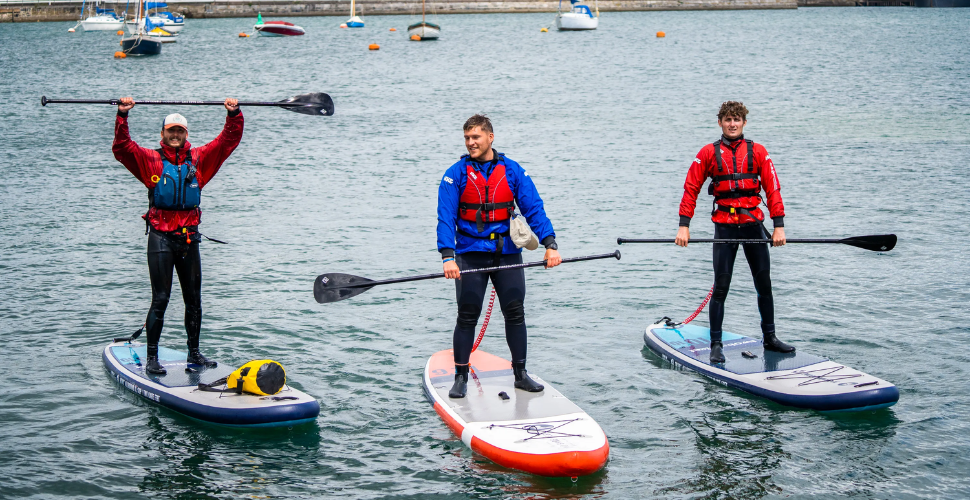 Three people standing on paddleboards raising the paddles above their heads with boats in the background